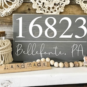 May include: Gray wooden sign with white painted text. The sign reads "16823 Bellefonte, PA". The sign is sitting on a white surface with wooden beads and a small sign that reads "handmade".