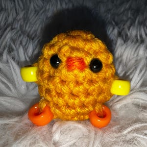 Puede incluir: Un juguete de pollito amarillo tejido a crochet con ruedas de plástico naranja y amarillo. El pollito tiene ojos negros y un pico rojo.
