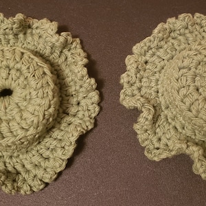 May include: Two handmade, sage green crochet coasters. Each coaster features a central, raised circle with a hole, surrounded by a ruffled edge. The coasters are displayed on a dark brown surface.