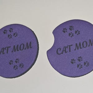 May include: Two purple round coasters with a black outline. Each coaster has the text "CAT MOM" and several black paw prints.