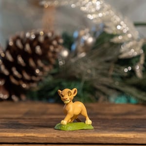 May include: A small, yellow lion figurine with a green base. The figurine is standing on a wooden surface.