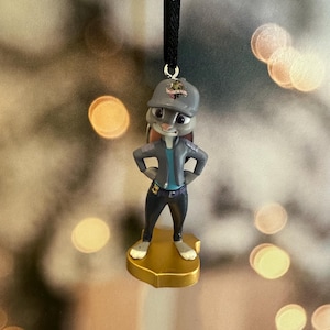 May include: A small, detailed figurine of a rabbit character wearing a gray cap and jacket, standing on a gold-colored base. The figurine has its arms on its hips and is suspended by a black cord, suggesting it's an ornament.