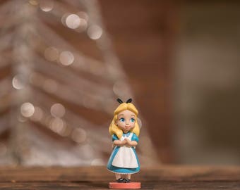 以下が含まれることがあります： アリス・イン・ワンダーランドの小さな青と白の置物。青いドレスに白いエプロンを着て、金髪に黒いリボンをしています。赤い台座の上に立っています。