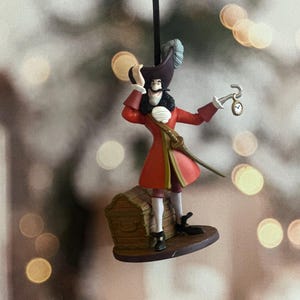 May include: Ornament of Captain Hook from Peter Pan. The figure wears a red coat, black boots, and a tricorn hat with a feather. He holds a hook and a pocket watch. The ornament is attached to a black string.