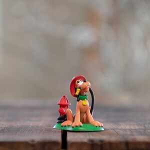 May include: A small plastic figurine of a cartoon dog wearing a red firefighter hat and holding a hose. The dog is standing on a green base with a red fire hydrant.