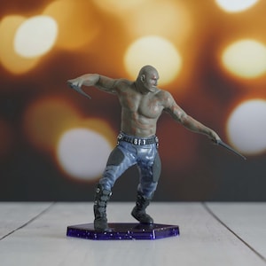 May include: A green and gray action figure of a character with a muscular build, wearing blue pants and a black belt. The figure is holding two black weapons and is standing on a purple base.