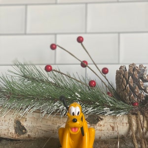 Puede incluir: Una figurita de plástico amarilla de Pluto, el perro de Disney, sentado sobre una base negra. La figurita está rodeada de una rama de pino verde con bayas rojas y una piña marrón.