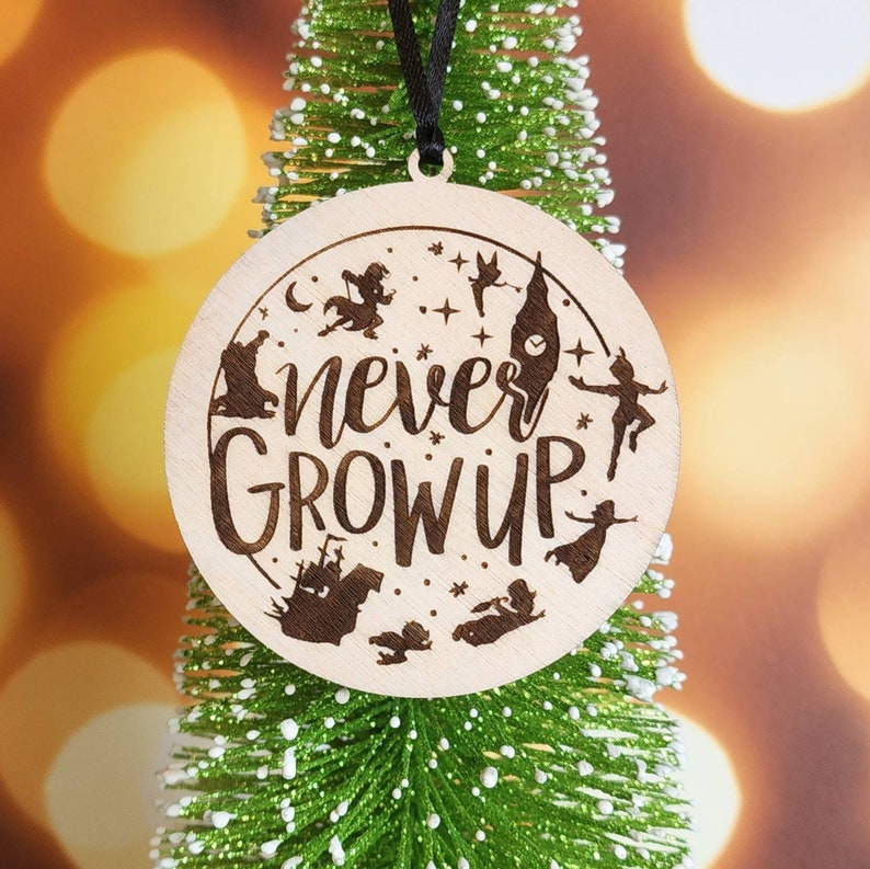 以下が含まれることがあります： 茶色の背景に黒い文字で「never grow up」と書かれた木製クリスマスオーナメント。オーナメントには、ピーターパン、フック船長、ティンカーベル、迷子の子供たちなど、映画「ピーターパン」のキャラクターのシルエットが描かれています。