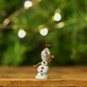 May include: A small white snowman figurine with brown twig arms and a carrot nose. The snowman is wearing a friendly smile and is standing on a wooden surface with a blurred green background.