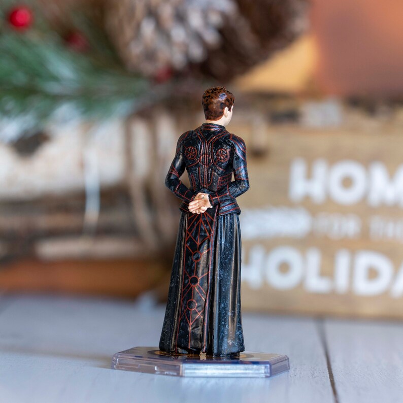 May include: A black and red detailed figurine of a person wearing a long robe. The figurine is standing on a clear plastic base.