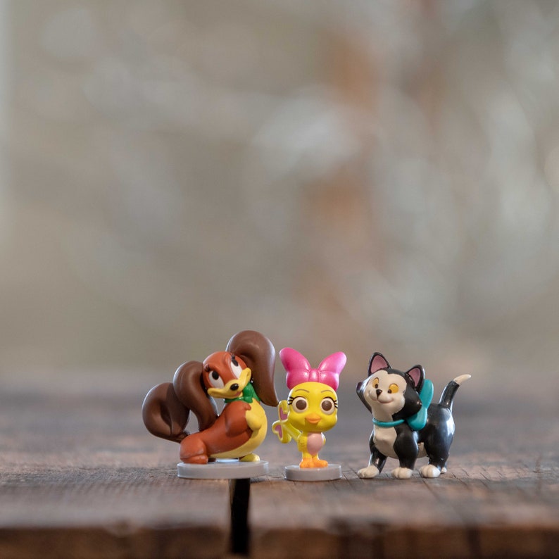 May include: Four small plastic figurines of cartoon characters. A brown and white dog with a green bow, a yellow bird with a pink bow, a yellow chick with a pink bow, and a black cat with a blue bow.