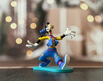 May include: A colorful figurine of Goofy in a dynamic pose, wearing a blue outfit with yellow and pink accents. The figure has white gloves and a black tail. It stands on a light blue base, perfect for Disney fans.