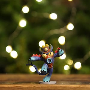 May include: A whimsical figurine of a cartoon monster in shades of blue, purple, and orange. The creature has three yellow eyes, horns, and a tail. It is posed in a dynamic stance on a small, round base.