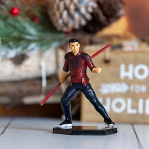 May include: A red and black action figure of a man holding a red staff. The figure is standing on a black base. The figure is posed in a fighting stance.
