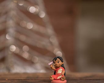 Peut inclure: Une petite figurine de Lilo du film Disney Lilo & Stitch. Elle porte une robe rouge et blanche et tient une fleur rose. La figurine est assise sur une base rose.
