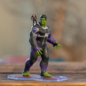 May include: A green and black action figure of the Hulk with a smaller action figure of Ant-Man standing on his shoulder. The Hulk is wearing a purple and black suit with a diamond pattern on the chest. The figures are standing on clear plastic bases.