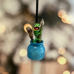 May include: A whimsical ornament featuring a green chameleon holding paintbrushes in a blue pot. The chameleon has a wide, open mouth. The ornament hangs from a black string, perfect for an artist or art lover.