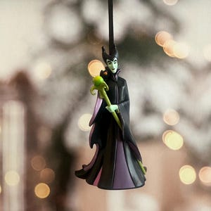 May include: A Maleficent Christmas ornament. The figure is wearing a black robe with purple accents and a green staff. The ornament is hanging from a black string. The background is blurred with bokeh lights.