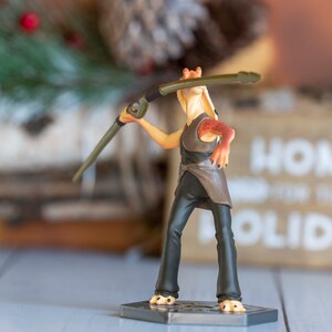 May include: A brown and orange action figure of a character from Star Wars holding a weapon. The figure is standing on a black base with a silver design.