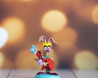 May include: A White Rabbit figurine from Alice in Wonderland. The rabbit wears a red coat and holds a teacup and saucer. The figurine stands on a light blue base, set against a blurred, warm-toned background.