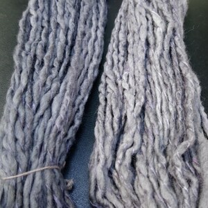 K&ouml;nnte beinhalten: Zwei Kn&auml;uel handgesponnener Wolle in hellgrau mit einem dezenten violetten Farbton. Die Wolle besteht aus Naturfasern und hat einen rustikalen, handgefertigten Look.