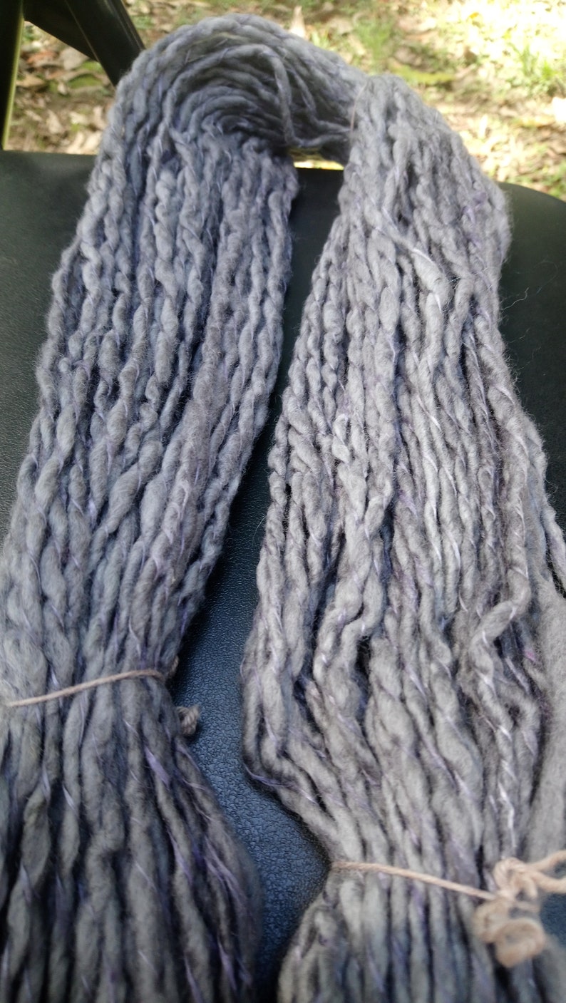 K&ouml;nnte beinhalten: Zwei Kn&auml;uel handgesponnener Wolle in hellgrau mit einem dezenten violetten Farbton. Die Wolle besteht aus Naturfasern und hat einen rustikalen, handgefertigten Look.