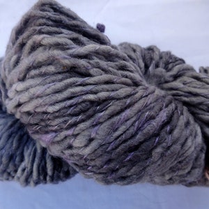 K&ouml;nnte beinhalten: Eine Kn&auml;uel dickes Garn in dunkelgrau mit violetten Akzenten. Das Garn ist aus Wolle und perfekt zum Stricken oder H&auml;keln.