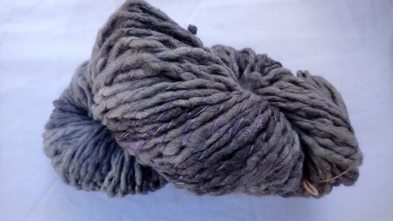 K&ouml;nnte beinhalten: Eine Kn&auml;uel dickes Garn in dunkelgrau mit violetten Akzenten. Das Garn ist aus Wolle und perfekt zum Stricken oder H&auml;keln.
