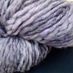 K&ouml;nnte beinhalten: Eine Kn&auml;uel dickes Garn in einer grauen und violetten Farbkombination. Das Garn ist weich und flauschig und hat einen dezenten Glanz.
