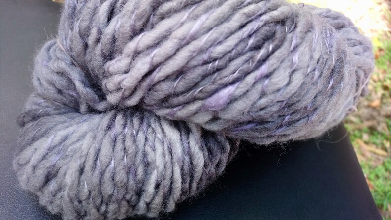 K&ouml;nnte beinhalten: Eine Kn&auml;uel dickes Garn in einer grauen und violetten Farbkombination. Das Garn ist weich und flauschig und hat einen dezenten Glanz.