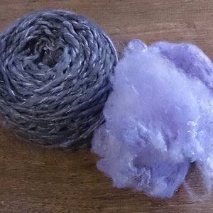 K&ouml;nnte beinhalten: Eine Kugel silbernes Garn mit einer violetten, flauschigen Faser daneben.