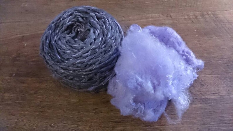 K&ouml;nnte beinhalten: Eine Kugel silbernes Garn mit einer violetten, flauschigen Faser daneben.