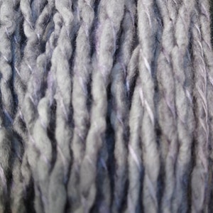 K&ouml;nnte beinhalten: Nahaufnahme eines dicken, grauen Garns mit einem dezenten violetten Schimmer. Das Garn ist gedreht und texturiert und erzeugt so einen einzigartigen und interessanten Look.