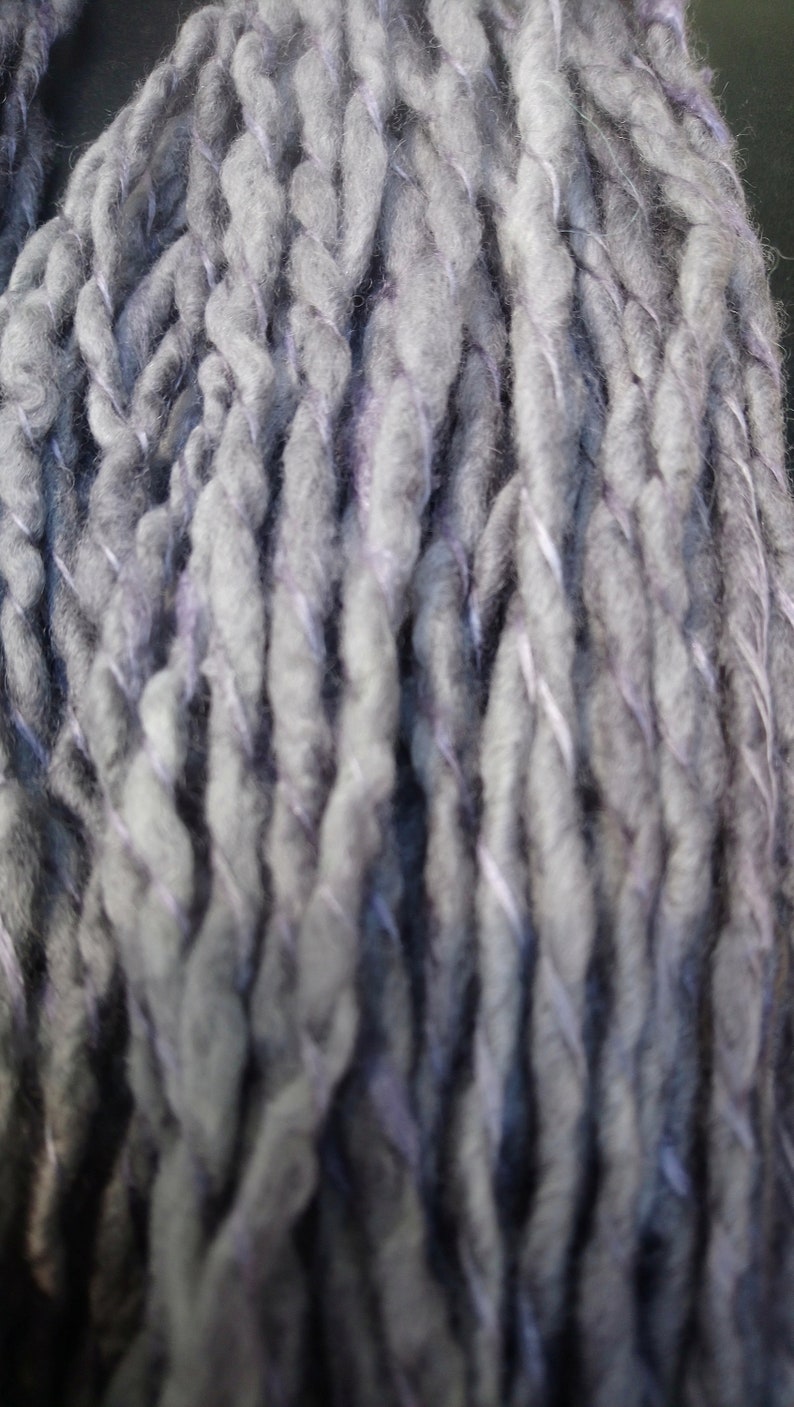 K&ouml;nnte beinhalten: Nahaufnahme eines dicken, grauen Garns mit einem dezenten violetten Schimmer. Das Garn ist gedreht und texturiert und erzeugt so einen einzigartigen und interessanten Look.