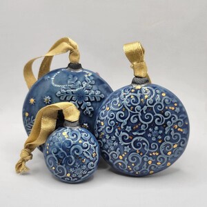 Peut inclure: Trois boules de Noël bleues en céramique avec des accents dorés et des rubans dorés. Les boules présentent des motifs de flocons de neige et de volutes. La plus grande boule mesure environ 10 cm de diamètre, la moyenne 7,6 cm et la plus petite 5 cm.