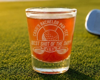 May include: A clear shot glass filled with amber liquid, featuring the text "Craig's Bachelor Par-Tee" and "Best Shot of the Day!" with a golf ball and club design. The glass sits on green grass with a golf ball and club in the background.