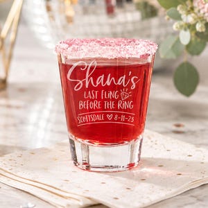 Puede incluir: Un vaso de chupito transparente con borde de az&uacute;car rosa, lleno de l&iacute;quido rojo. El vaso est&aacute; grabado con "Shana's Last Fling Before The Ring Scottsdale 8-11-23". El vaso est&aacute; sobre una servilleta blanca con motas doradas.