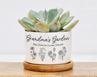 May include: White ceramic planter with the text "Grandma's Garden" and the names Max, Emmie, Lucas, and Garrett. The planter has a black outline of four different flowers. A succulent plant is in the planter.