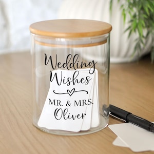 May include: A clear glass jar with a wooden lid, labeled "Wedding Wishes" with a heart and "MR. & MRS. Oliver." The jar contains white cards and sits on a wooden surface next to a black pen and two blank cards. Green foliage is in the background.