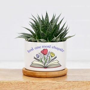 May include: White ceramic planter with a book and flowers design. The text on the planter reads "Just one more chapter". A succulent plant is growing in the planter.