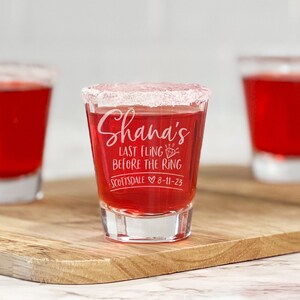 Puede incluir: Tres vasos de chupito llenos de l&iacute;quido rojo, uno enfocado con el texto "Shana's Last Fling Before the Ring Scottsdale 8-11-23". El borde del vaso central est&aacute; recubierto con una sustancia blanca, probablemente sal o az&uacute;car. Los vasos est&aacute;n sobre una tabla de madera.