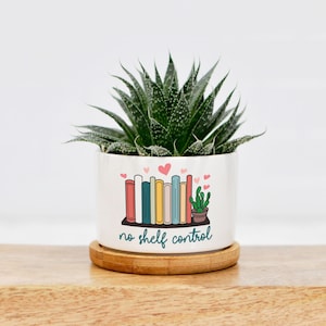 May include: White ceramic planter with a green succulent plant. The planter has a colorful illustration of books on a shelf with the text "no shelf control".