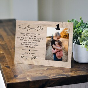 May include: A wooden photo frame with a clip at the top. The frame has a printed message that reads "To our Bonus Dad. Thank you for loving us as your own. You may not have our genes, but you will forever have our heart. Not half. Not step. Just family. Love always. Emmy + Sutton". The frame has a photo of two children hugging.