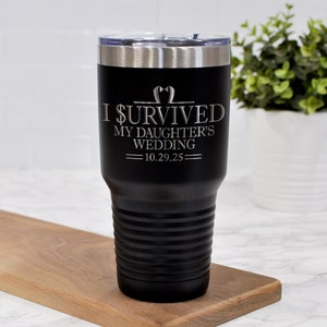 May include: Black insulated tumbler with a silver rim and lid. The tumbler has the text "I SURVIVED MY DAUGHTER'S WEDDING 10.29.25" in silver lettering. The tumbler is sitting on a wooden cutting board.