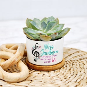 May include: A white ceramic succulent planter with a bamboo base. The planter has a musical note design and the text "Mrs. Jacobson without you our class would Bb".