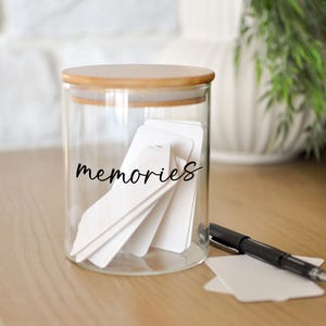 May include: A clear glass jar with a wooden lid, filled with white cards. The word "memories" is written in black script on the jar. A black pen and additional white cards are on the wooden surface next to the jar. Green foliage is in the background.