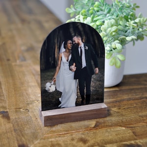 Puede incluir: Un soporte de fotos de madera con la parte superior redondeada, que muestra una foto en blanco y negro de una pareja que se toma de la mano. La pareja est&aacute; vestida con atuendos formales, la mujer con un vestido blanco y el hombre con un traje.
