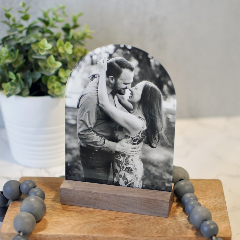 Puede incluir: Una foto en blanco y negro de una pareja abraz&aacute;ndose, impresa en un arco de acr&iacute;lico transparente, exhibida sobre una base de madera.