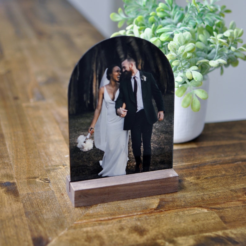 Puede incluir: Un soporte de fotos de madera con la parte superior redondeada, que muestra una foto en blanco y negro de una pareja que se toma de la mano. La pareja est&aacute; vestida con atuendos formales, la mujer con un vestido blanco y el hombre con un traje.