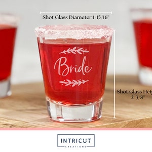 Puede incluir: Un vaso de chupito transparente lleno de l&iacute;quido rojo, con borde blanco y grabado con la palabra "Bride" y detalles florales. El vaso de chupito mide 6 cm de alto y 5 cm de di&aacute;metro. El vaso de chupito est&aacute; sobre una superficie de madera.